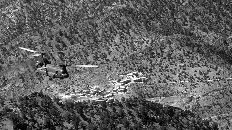 Birds eye view of a tandem rotor helicopter flying over a rocky mountain terrain.