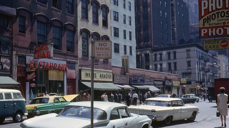 City street filled with cars in front of a row of residential buildings with local storefronts on a sunny day.