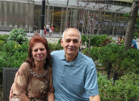 A gray-haired man (right) in a light blue top sits with his arm around a red-headed woman, with greenery in the background.