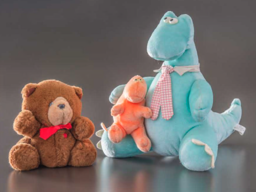 A brown stuffed bear wearing a red tie (left) and a light blue stuffed dinosaur wearing a pink tie, holding a smaller orange stuffed dinosaur